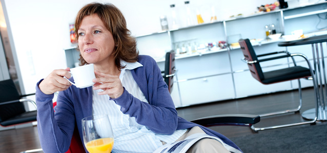 Frau trinkt entspannt aus einer Tasse, vor ihr ein Glas Orangensaft